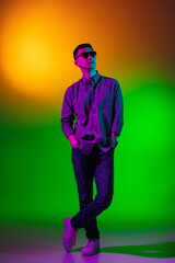 Young man, fashion model in casual clothes isolated on studio background in gradient green yellow neon light, colour filter. Concept of human emotions, facial expression.