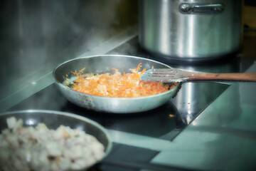 Close up of frying pan with fresh vegetables and wooden spatula. Kitchen is equipped with induction cookers