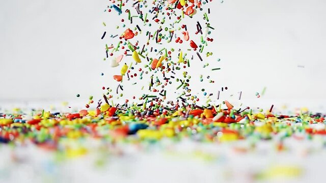 Multi-colored differently shaped sweet sprinkles are poured on a wooden white table on a white background. Slow motion bright fun video.