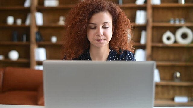Confident curly red-haired businesswoman using laptop in the office is solving difficult tasks, concentrated woman making some important desision, looks at the computer screen with puzzled face