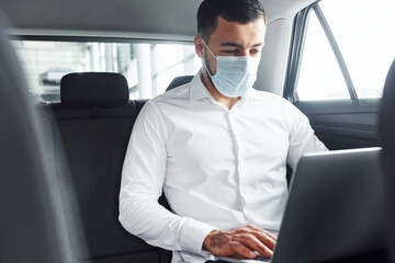 Works by using laptop. Young man in white shirt is sitting inside of a modern new automobile