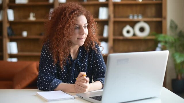 Young curly female student watching educational webinar and taking notes, sitting at the the desk and talking online, takes a part in virtual conference, discussing online and writes down
