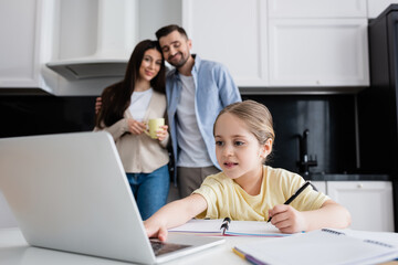 kid using laptop while doing homework near parents smiling on blurred background