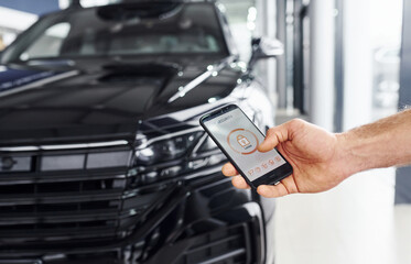 Fototapeta premium Black car. Close up view of man's hand that holds phone with labels and icons. Conception of remote control