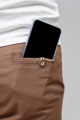 Close up of mobile phone with empty black screen in back pocket of brown pants on gray background, copy space.