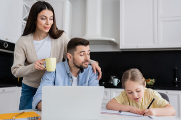 girl writing in notebook while doing homework near dad and smiling mother