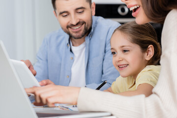 blurred woman pointing at laptop near daughter and smiling husband