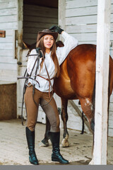 Young woman rider wearing a white shirt and hat with her brown horse in a stall, portrait.