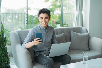Smiling young man typing on mobile phone while sitting on a sofa at home with laptop computer