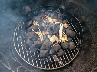 Glowing charcoal briquettes with smoking wood on a charcoal grate in a kettle grill during grilling.