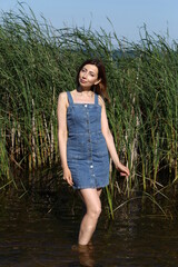 A slender woman in a denim dress standing in the water against the background of reeds