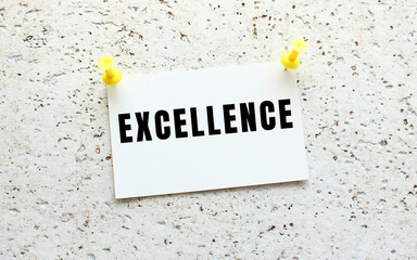 EXCELLENCE is written on a card attached to a white textured wall with a button. Office reminder.