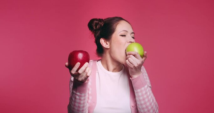 Beautiful young caucasian girl holds two apples, red and green in her hands and funny bites quickly each one in turn isolated on pibk background. Vegetarian healthy fruits. Problem of choice concept