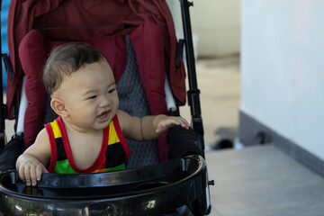 A 6 month old boy wearing a colorful shirt sits in a red stroller and opens his mouth to call his parents.