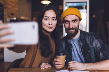 Portrait of good looking male and female friends sitting at cafeteria table and posing while clicking smartphone selfie pictures, millennial hipster guys looking at camera and photographing