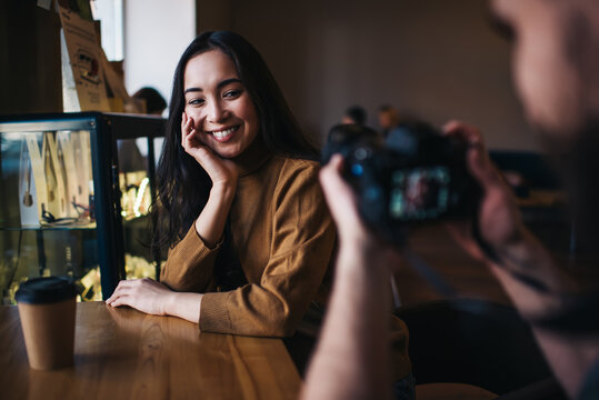 Back view of male photographer using modern technology for taking pictures of pretty Kazakh woman, amateur making photos of cheerful hipster girl sitting at cafeteria table and smiling at camera