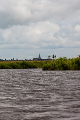 Le village de Saint Joachim photographié depuis les marais avec un ciel couvert. L´eau du marais clapote et les roseaux sont jaunes et verts. L´église de ce village est visible au loin.