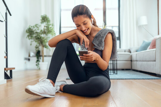 Sporty Young Woman Using Her Mobile Phone While Holding Water Bottle After A Pilates Class On The Floor At Home.