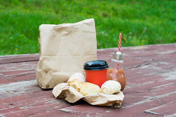 Juice or smoothie in a glass bottle with a straw, coffee and sweet buns with jam on a wooden table. Paper cup and paper bags for take-away food. The concept of outdoor recreation.