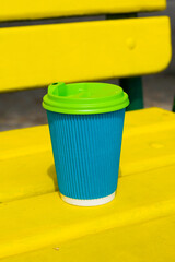 Paper cup blue color with a coffee to go on yellow wooden bench on the beach or in the park in the summer sunny day.