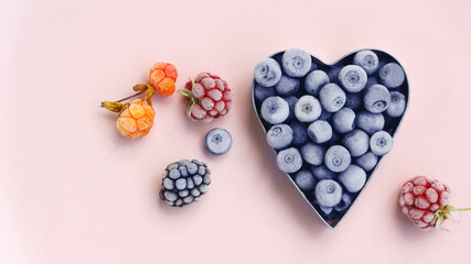 freshly frozen blueberries in the shape of a metal heart, raspberries, blackberries and cloudberries on a pink background. summer berry picking