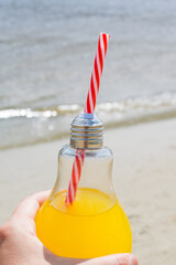 Fresh or cocktail in a glass bottle with a straw close-up in a female hand against the background of the beach and water, vertical format. Travel and recreation concept.