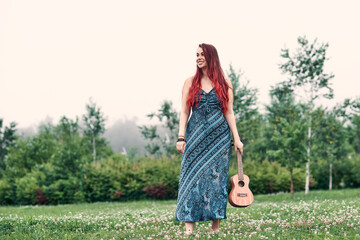 Smiling woman with red hair in a blue dress stands in the park on a summer day. Image in copy space