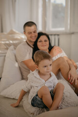 Fototapeta premium Front view of happy smiled boy with mother and father on background. Smiled parents hugging and small son playing, sitting on couch. Adopted child with lovely family. Concept of healthy relationship.