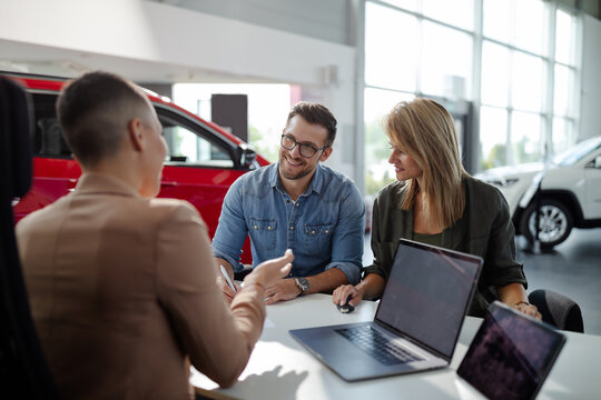 Smiling saleswoman talking with young couple about contract for new car in modern car showroom