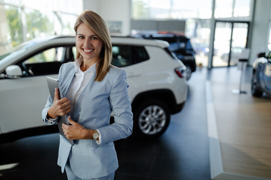 Female salesperson at a car showroom, holding a laptop and smiling to the camera.