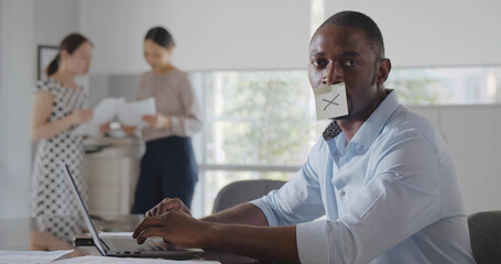 Businessman typing on laptop in office with sticker note with cross on his mouth