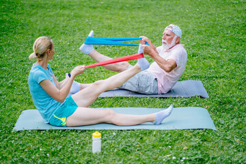 Senior sportsman and woman physiotherapist sitting on yoga mat in green grass meadow exercising with resistance band at park outdoor.