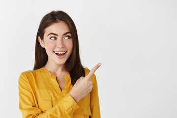 Close-up of lady smiling, pointing and looking right with surprised face, standing on white background