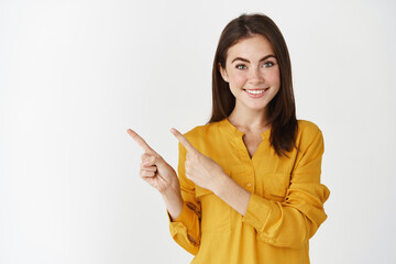 Close-up of young female model pointing fingers left at copy space, showing company logo and smiling, standing over white background - 444039478