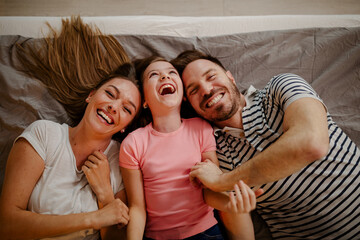 Beautiful young mother, father and their daughter looking at camera and smiling while lying on bed.
