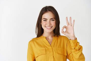 Young confident woman smiling, showing okay gesture, saying yes or alright and standing on white...