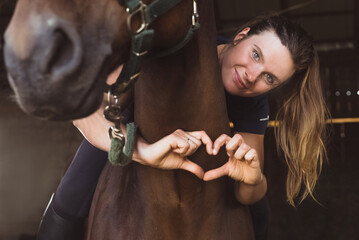 Horsewoman posing with her seal brown horse in the stable. Girl making a heart with her fingers....