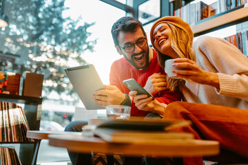 Close up photo of lovely young couple using modern technology, having coffee and fun while sitting in contemporary bookshop. Young couple in outfit with happy colors is laughing and having fun.