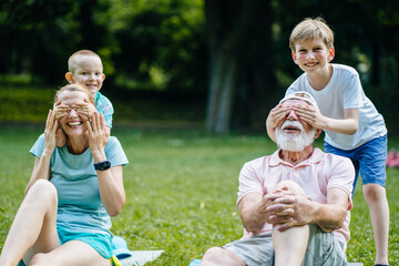 Guess who concept. Two funny brothers playing hide-and-seek game: sons close eyes with hands their mother and grandfather at the same time at green summer park outdoor.