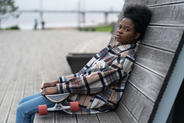Grumpy african young woman tired sit in modern urban space outdoors with long board in hands....