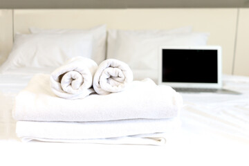 Stack of fresh white bath towels on bed sheet with as laptop computer blurred background., working remotely online on vacation in bedroom at hotel concept.