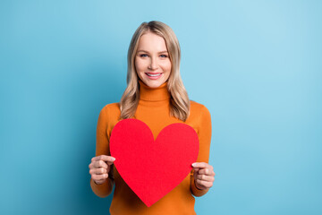 Photo of sweet adorable young woman dressed orange pullover smiling holding red heart isolated blue color background