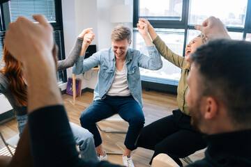 Group of happy young activity diverse multiethnic businesspeople holding hands raised, celebrate business success sitting in circle on background of window at modern office room in sunny day.