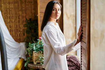 Half length portrait of Asian woman dressed in white shirt looking at camera during Sunday morning, attractive female posing during weekend leisure in home apartment or comfortable hotel room