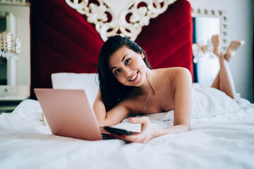 Portrait of happy Asian blogger with modern cellular and laptop technology smiling at camera during leisure time in hotel bedroom, carefree freelancer with digital smartphone and netbook resting