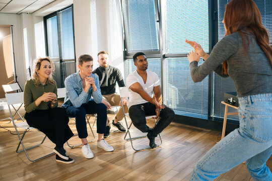 Happy diverse multi-ethnic office workers having fun playing in active games during team building in modern office room. Young happy woman playing charades showing pantomime to friends by window.