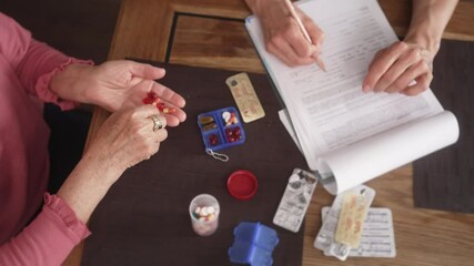 A top view of the elderly woman picking pills in the palm of her hand and a doctor writing down a medical history