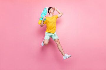 Full size photo of young guy happy positive smile jump look forward hold water pistol isolated over pink color background