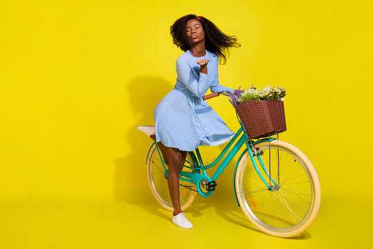 Full body photo of brunette afro american wear red headband blue dotted dress bike send air kiss isolated on yellow color background