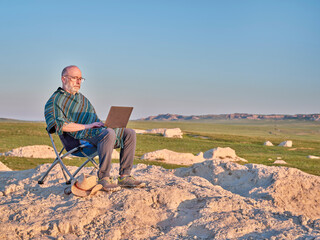 Senior man in Mexican poncho is sitting on a folding chair and working on laptop in the middle of...